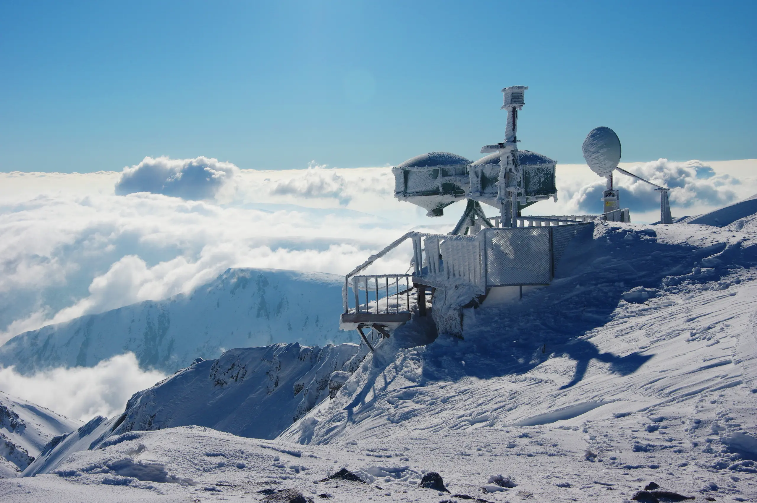 Snow-covered weather station on mountain summit with clouds.